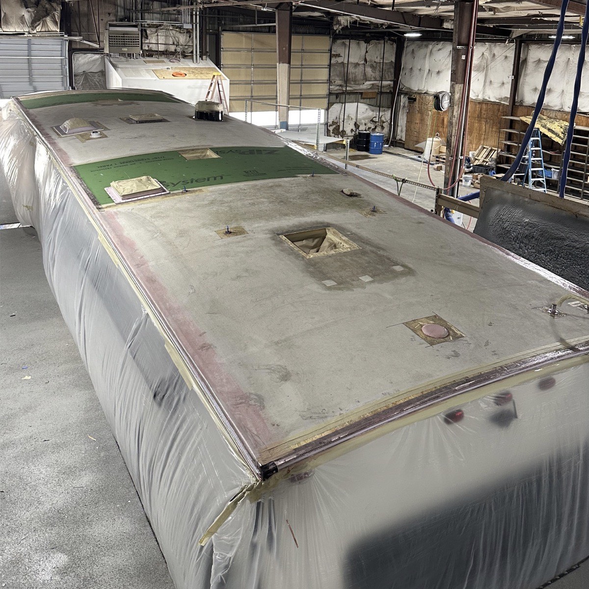 The same RV in the shop before spraying: the old membrane stripped back to bare, stained decking, vents and fixtures pulled, exposed sheathing at the front, and the sides masked off in plastic.